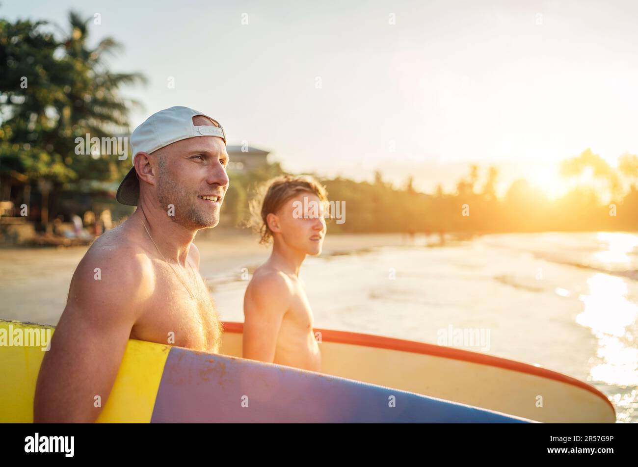 Lächelnder Mann mit jungendlichen Jungen Sohn mit Surfbrettern geht zum Surfen ins Meer. Sie haben einen Winterurlaub und genießen ein wunderschönes Sonnenuntergangslicht. Familie Stockfoto