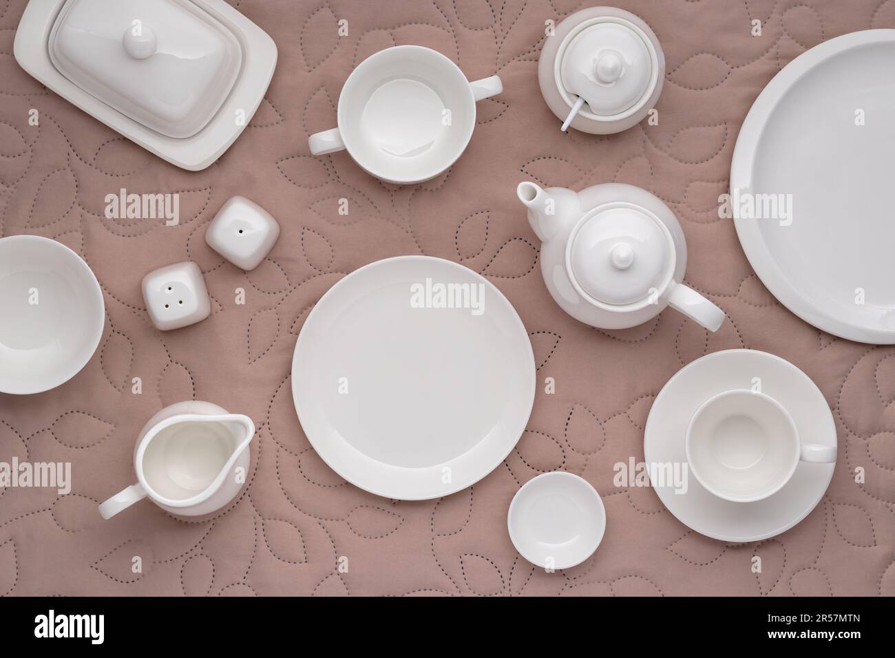 Weißes Porzellan-Geschirr zum Servieren und Essen von Mahlzeiten. Leere saubere Teller, Tasse, Teekanne, Zuckerdose, Milchkanne und Butterschale auf einem Tisch mit beigefarbenem fa Stockfoto