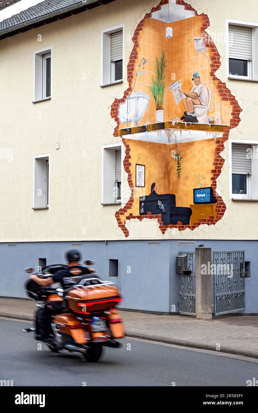 Haus mit lustigem Wandbild, Blick ins Innere, Mann liest Zeitung auf der Toilette, Frau im Wohnzimmer sieht sich den Tatort im Fernsehen an, Motorradfahrer, Biker an Stockfoto