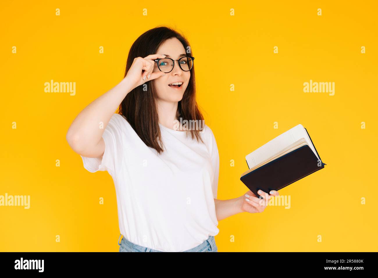 Porträt einer glücklichen jungen Frau, die eine Brille und ein weißes T-Shirt trägt, in ein Buch mit offenem Mund eingetaucht, isoliert auf gelbem Hintergrund. Stockfoto