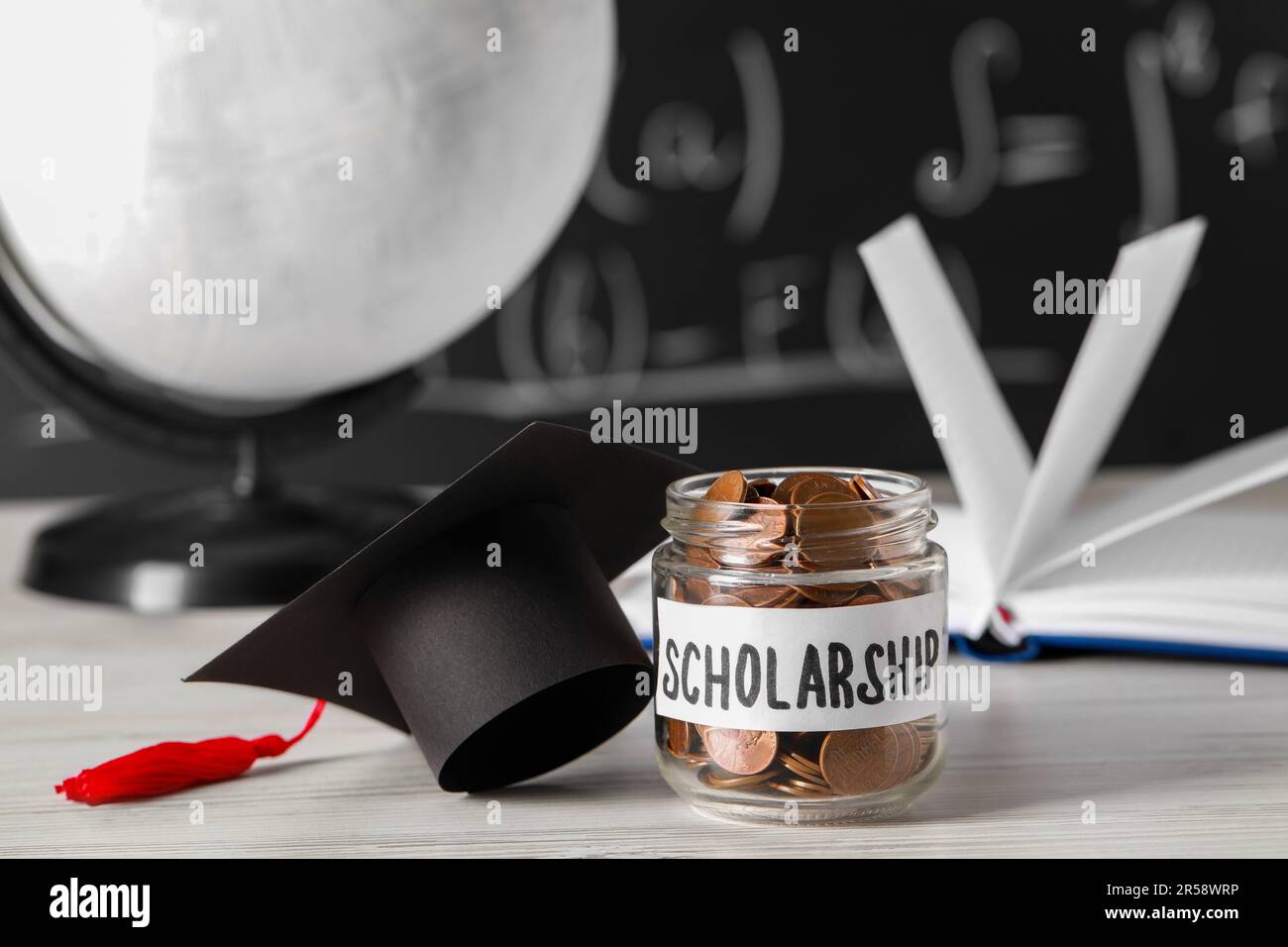 Stipendienkonzept. Glasbecher mit Münzen und Graduierungskappe auf weißem Holztisch Stockfoto