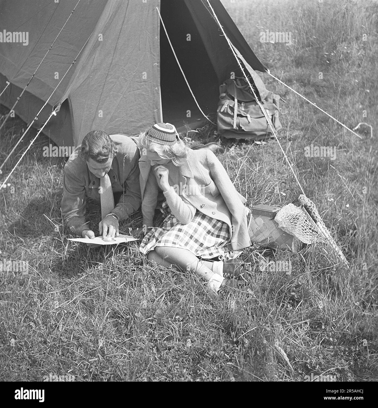 Camping in alten Zeiten. Das junge Paar Barbro Santesson und Kurt Ferström sitzen vor ihrem Zelt und studieren sorgfältig eine Karte und einen Kompass. Sie haben sich mit einem Rucksack und einem Fischernetz im Gras für das Leben im Freien ausgestattet. Aber sie scheinen nicht ganz hingebungsvoll zu sein, da er eine Krawatte und sie einen modischen Mantel trägt. Schweden 1945. Kristoffersson Ref. P45-5 Stockfoto