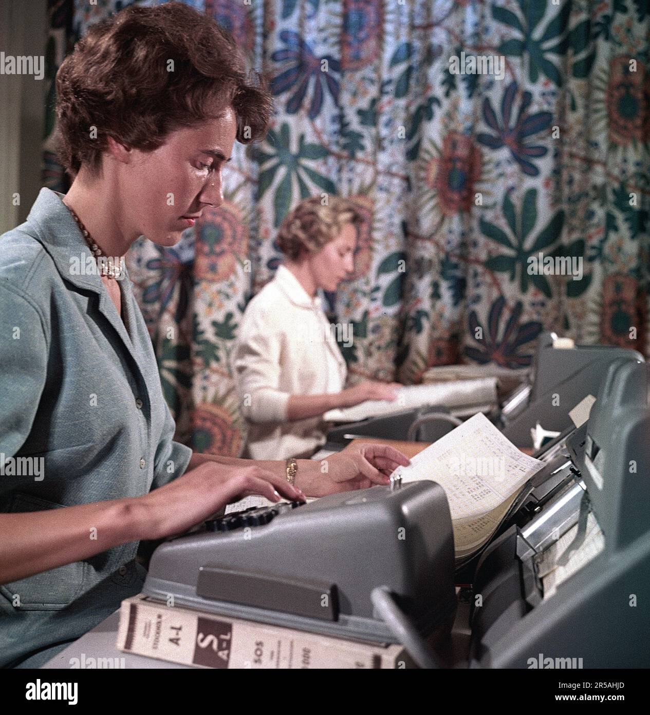 Büro in den 1950er. Eine Angestellte sitzt und zählt auf einen Taschenrechner. Andere Bürogeräte sind auch auf dem Desktop sichtbar. Ein Kollege sitzt im Hintergrund und führt die gleiche Art von Arbeit aus. Schweden 1958. ref. BV70-2 Stockfoto