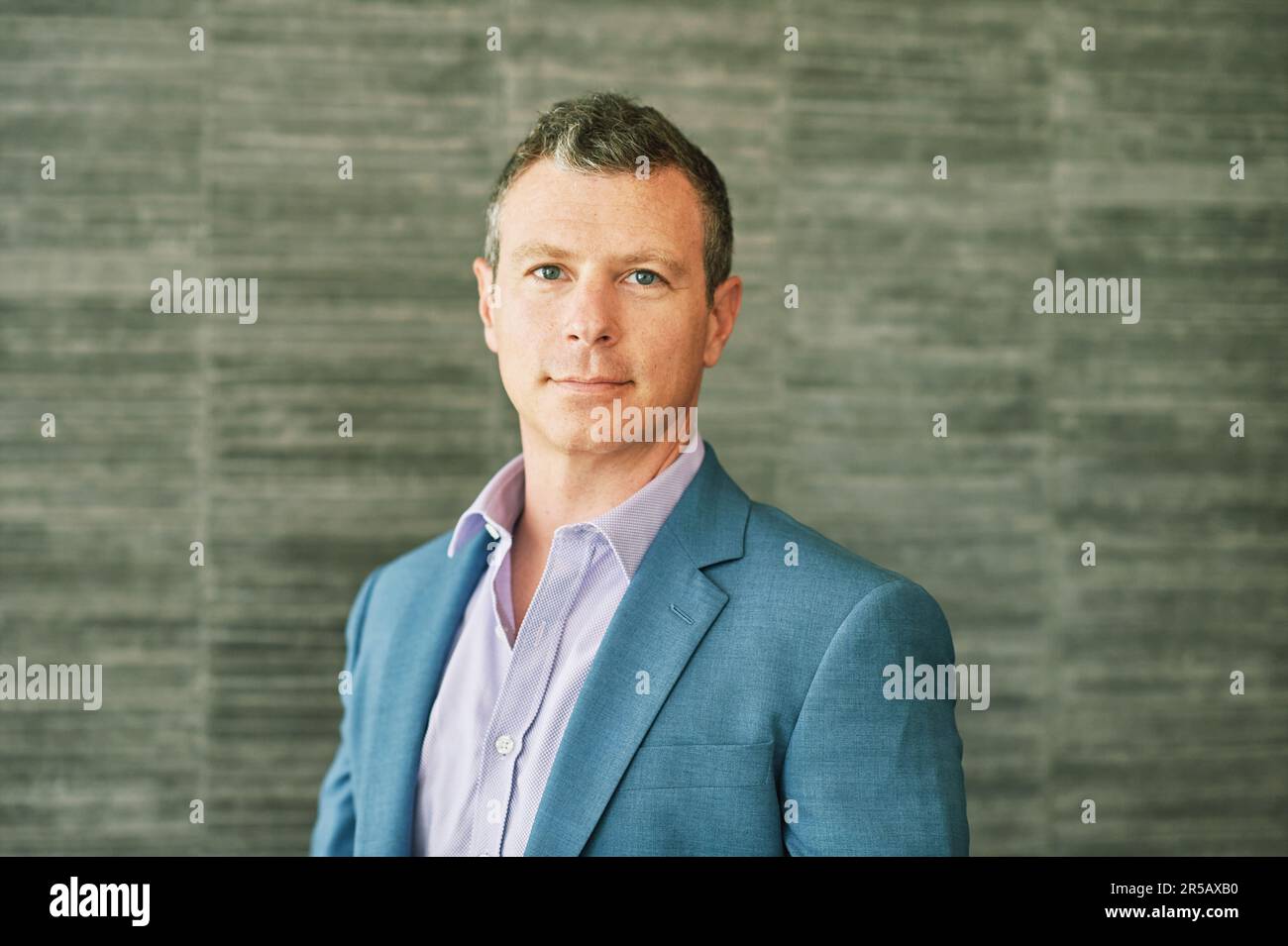 Porträt eines erwachsenen 40-jährigen Mannes, der vor grauem Hintergrund posiert, ein lila Hemd und eine blaue Jacke trägt Stockfoto