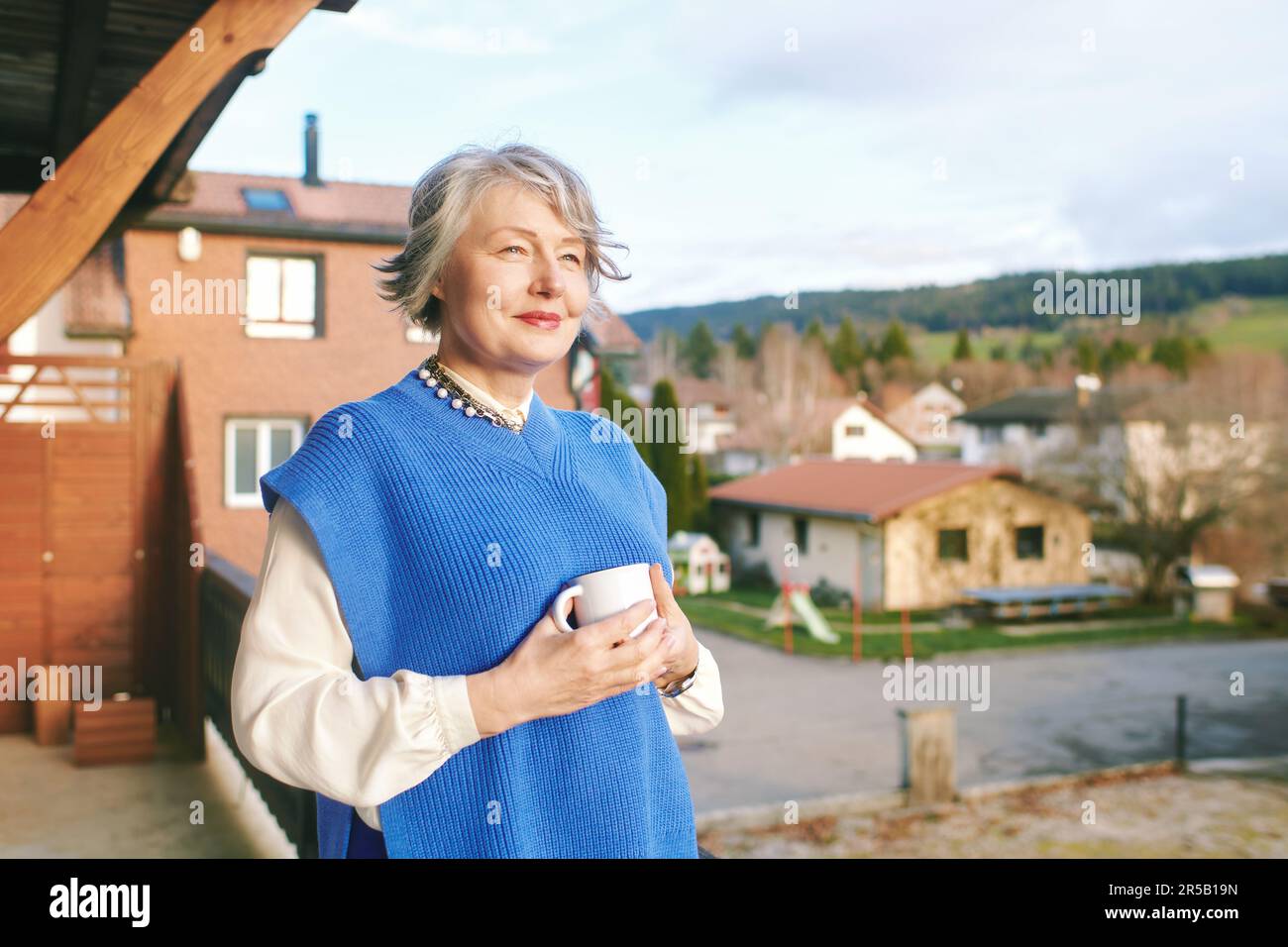 Porträt einer wunderschönen 50 -55-jährigen Frau, die auf dem Balkon posiert und eine Tasse Tee oder Kaffee hält Stockfoto