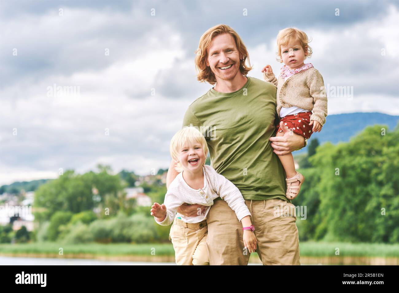 Außenporträt eines glücklichen jungen Vaters, der zwei Kinder in den Armen hält, Kleinkinder und Vorschulkinder, die an einem schönen sonnigen Tag Spaß mit ihrem Vater haben Stockfoto