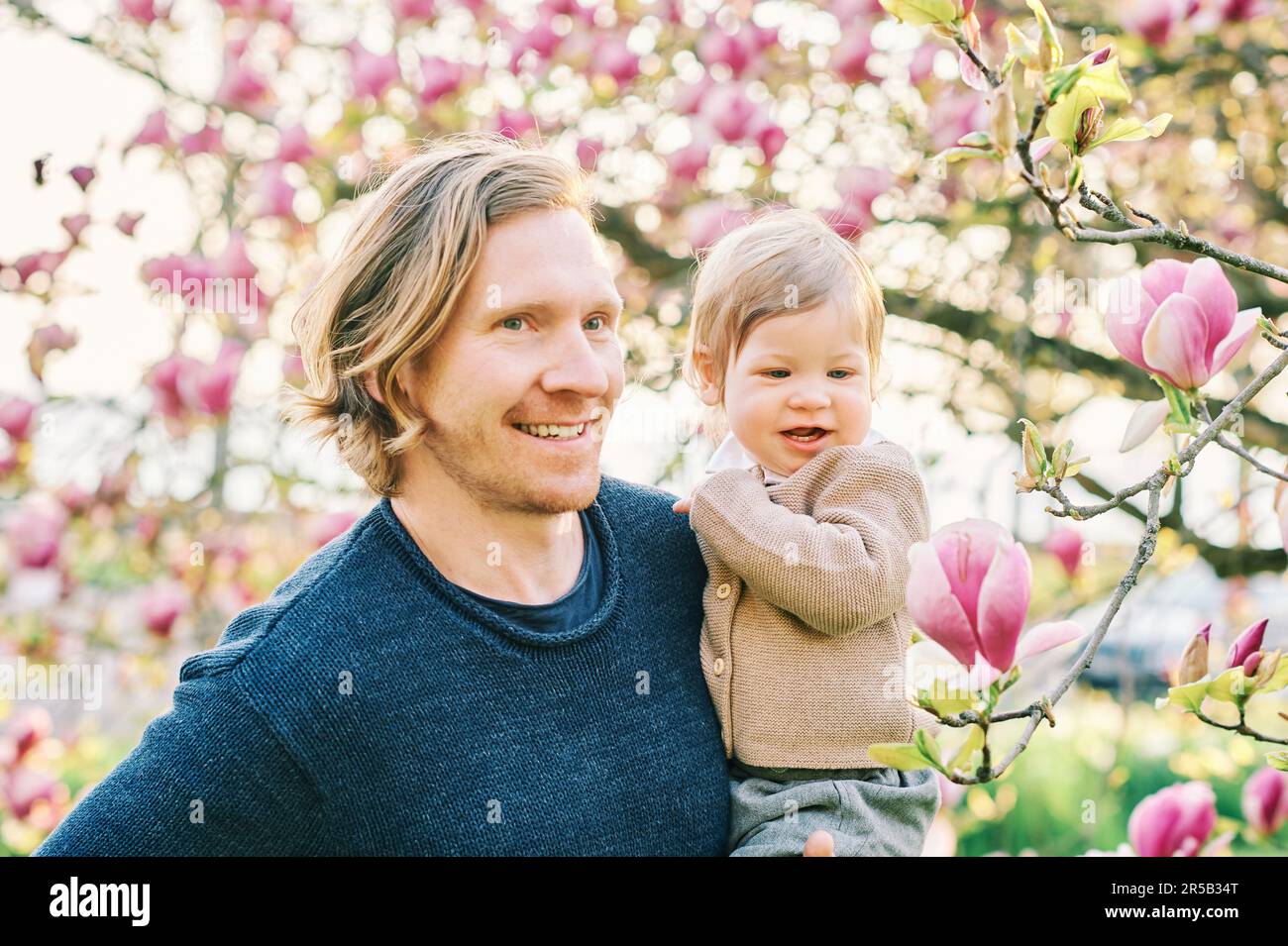 Außenporträt eines glücklichen jungen Vaters, der im Frühlingspark mit einem niedlichen Kleinkind spielt und sich wunderschöne blühende Magnolienblüten anschaut Stockfoto