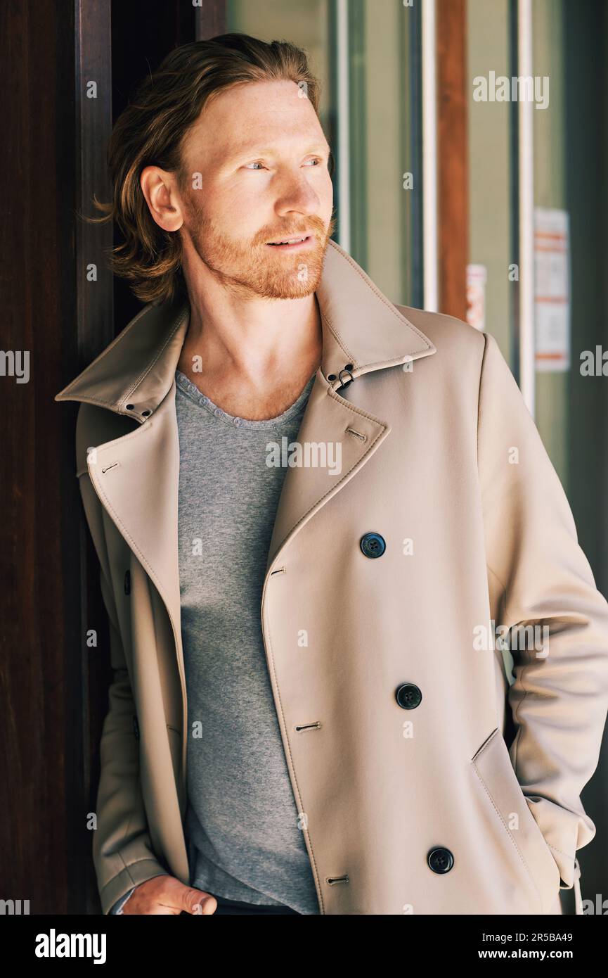 Außenporträt eines attraktiven jungen Mannes mit rotem Haar, beigefarbenem Mantel, der sich an die Fassade lehnt Stockfoto
