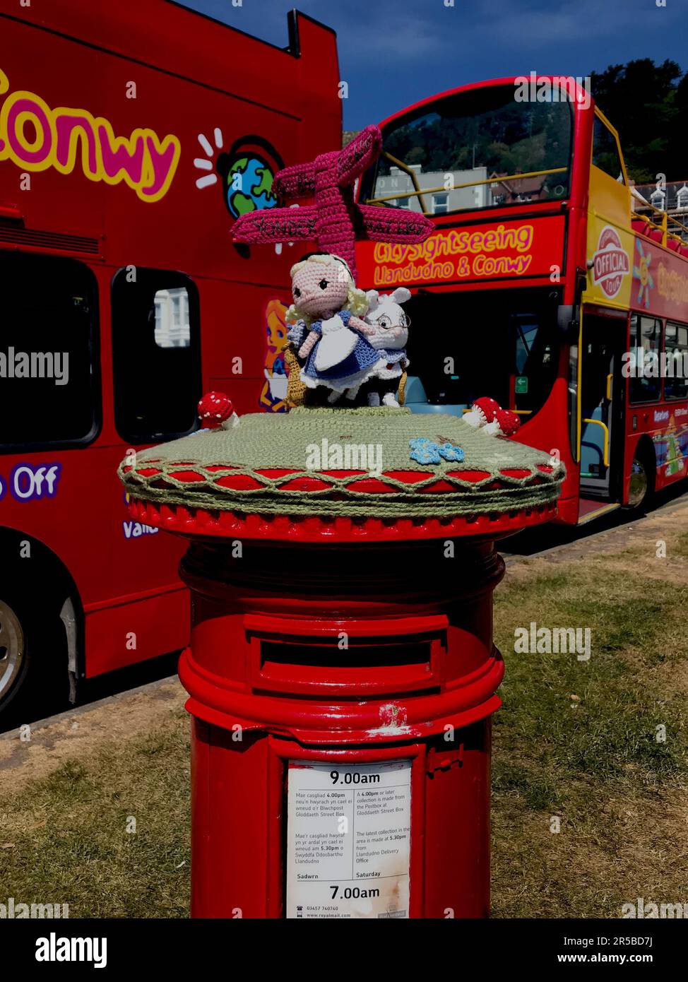 Briefkastenstricken. Alice im Wunderland thematisch gestrickte Hülle für die Oberseite des Postfachs, Alice thematisierte Tourbusse für Conwy und Llandudno im Hintergrund. Stockfoto