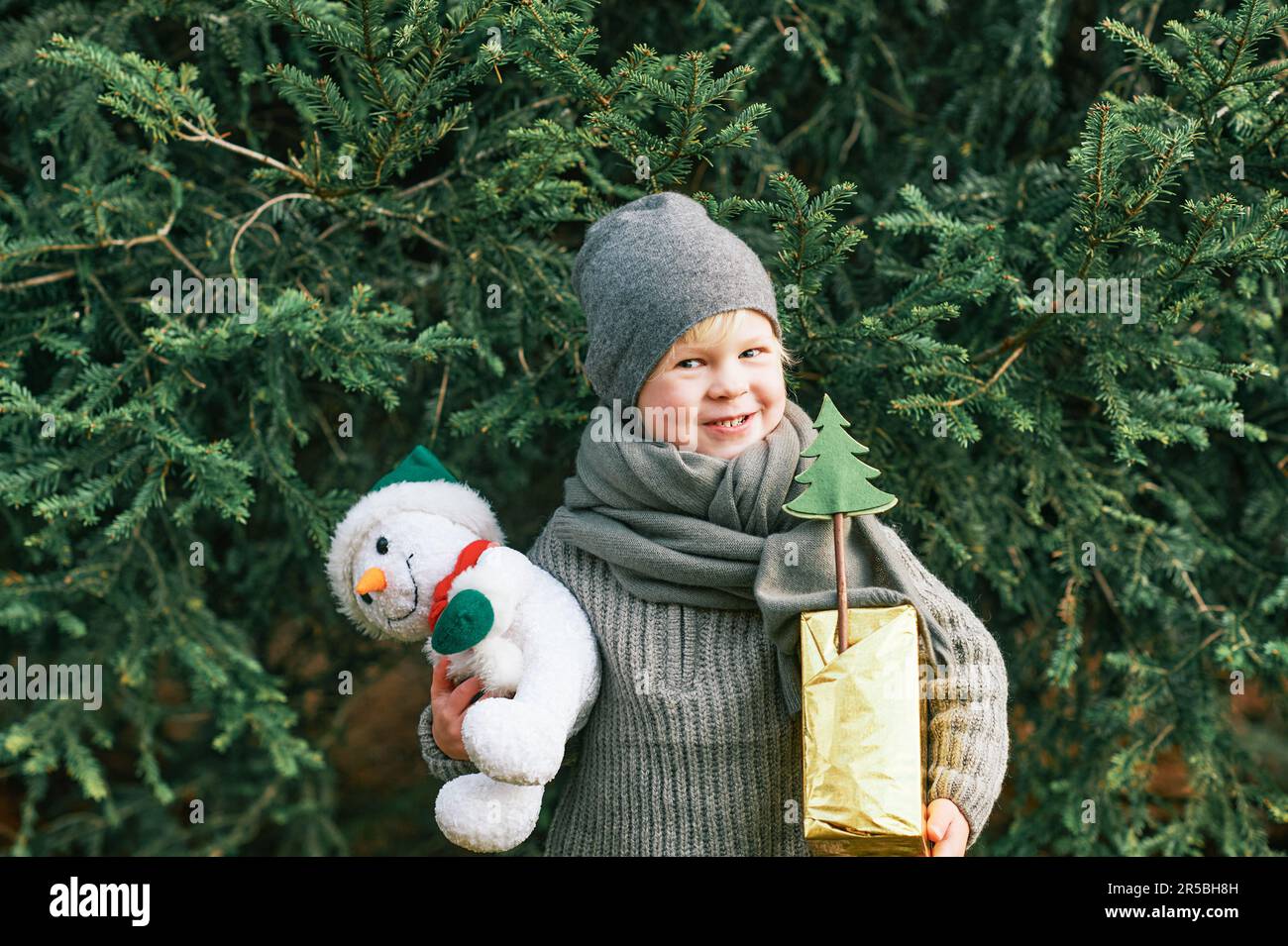 Außenporträt eines fröhlichen, süßen Jungen, der neben einer Kiefer posiert, eine Geschenkbox und ein Schneemannspielzeug in der Hand hält, einen Wurmschal und einen Hut trägt Stockfoto