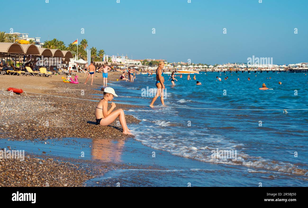 Antalya; Türkei-September 16; 2022: Ein kleines Mädchen, das auf den Kieseln am Meer sitzt. Leute sonnen sich, schwimmen oder laufen am Strand in summ Stockfoto