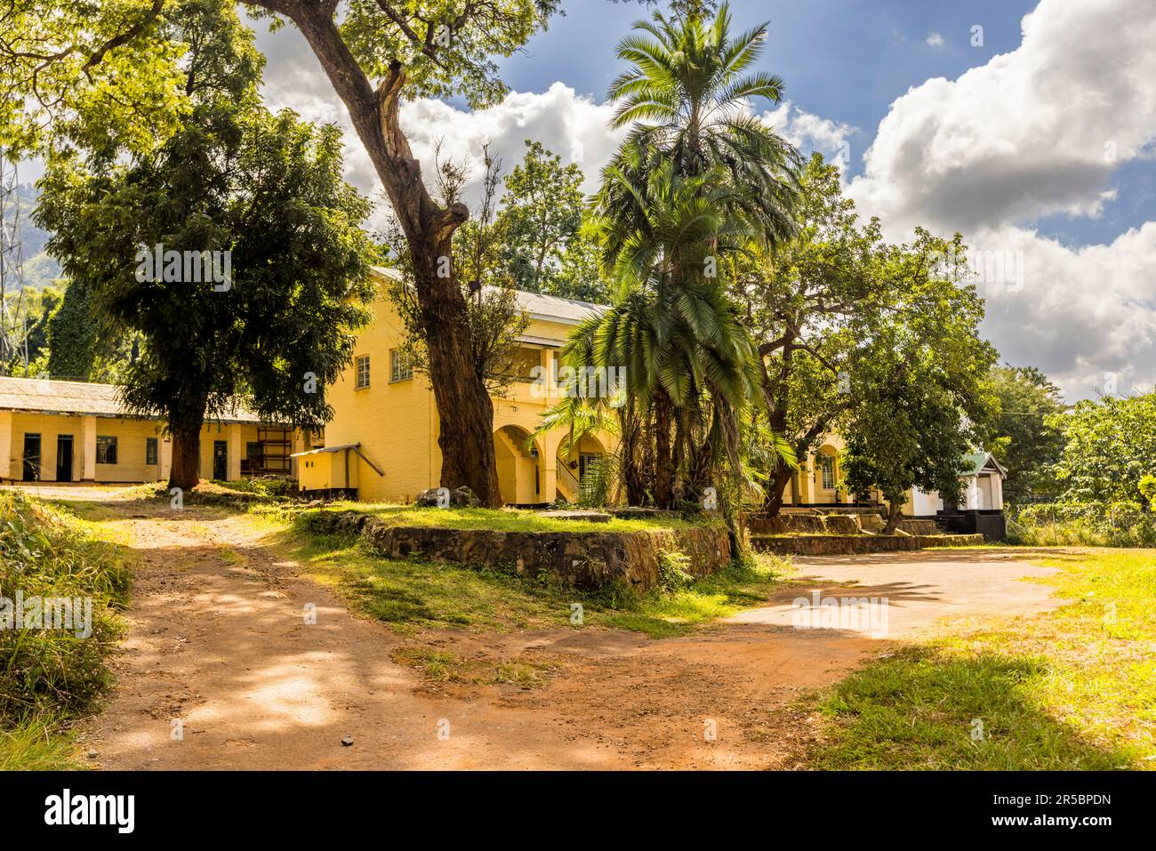 Ein cremefarbenes Kolonialgebäude mit gewölbten Arkaden steht unter alten Bäumen und Palmen auf dem Gelände der Geologischen Landesaufnahme in Zomba, Malawi. Zomba war von 1891 bis 1974 die Hauptstadt der britischen Kolonie Njassaland und später von Malawi, und sein Parlament tagte bis 1994 weiterhin in der Stadt. Stockfoto