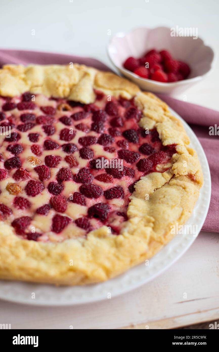 Hausgemachter Himbeerkuchen oder Galette auf weißem Holztisch über einem pinkfarbenen Tuch, mit frischen Beeren an der Seite, Seitenansicht, Nahaufnahme, vertikaler Shot. Stockfoto