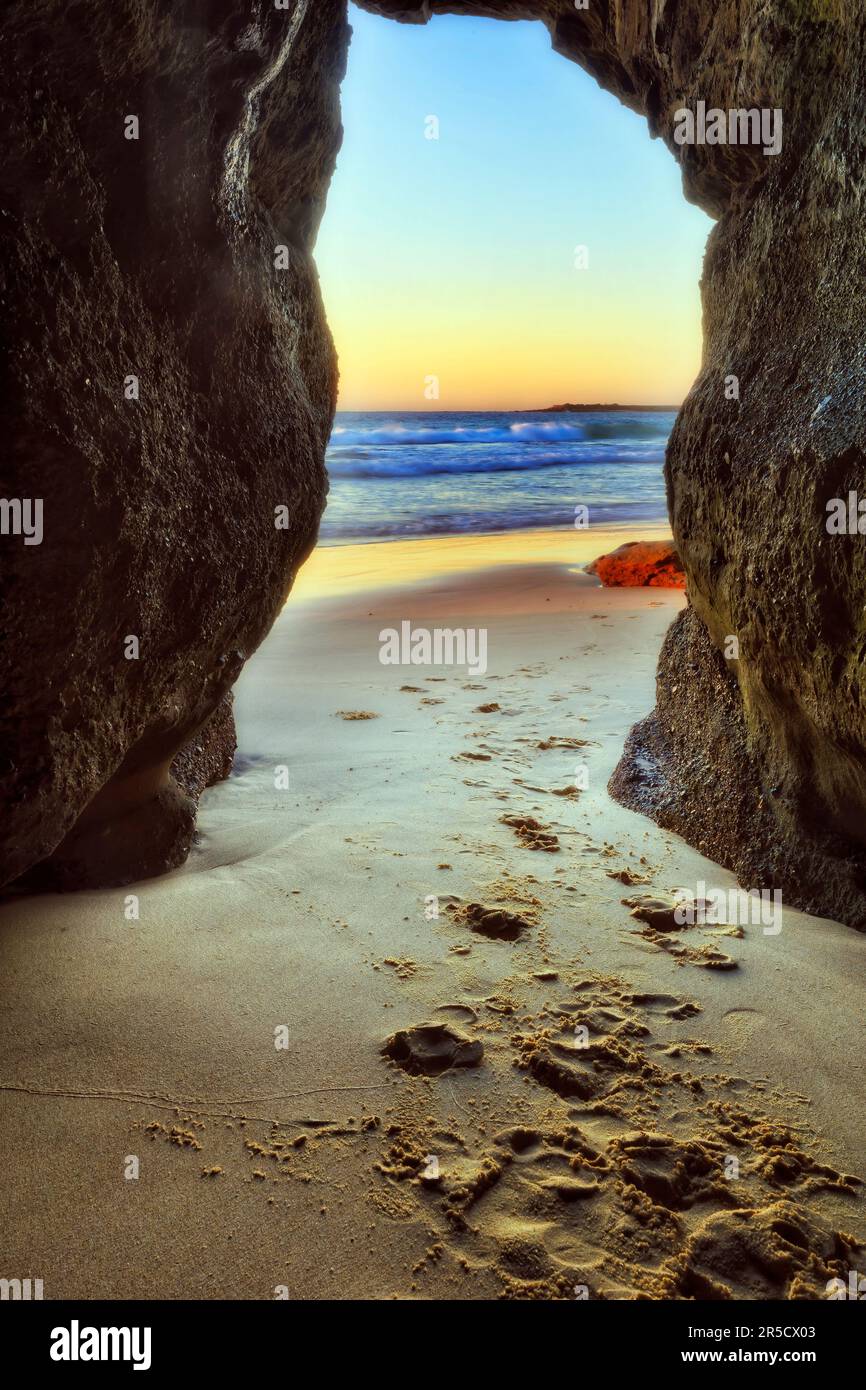 Fußspuren auf Sand, die während des Sonnenaufgangs durch eine Meereshöhle am Caves Beach in Australien zum Pazifischen Ozean führen. Stockfoto