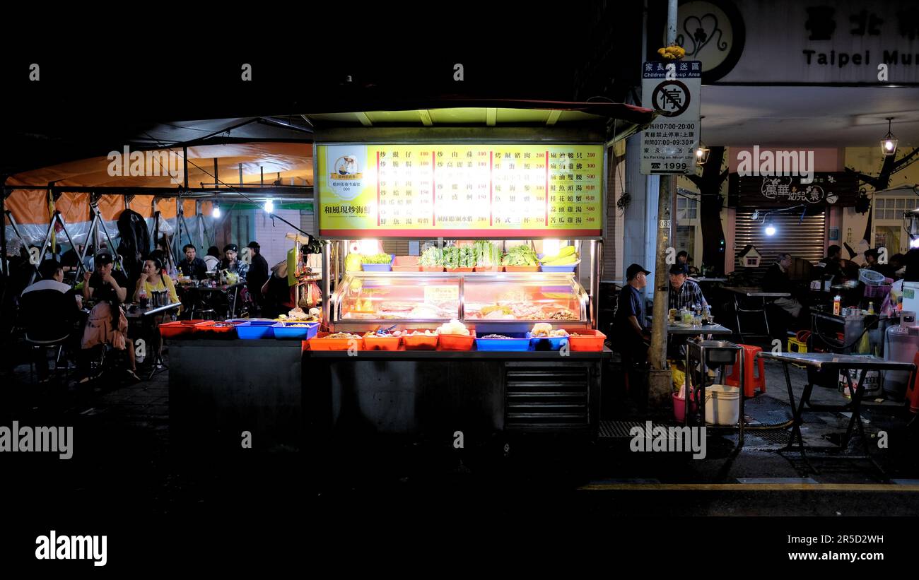 Street Food-Stand auf dem Huaxi Street Tourist Night Market in Taipei, Taiwan; Tische und Restaurantgäste sitzen im Hintergrund. Stockfoto