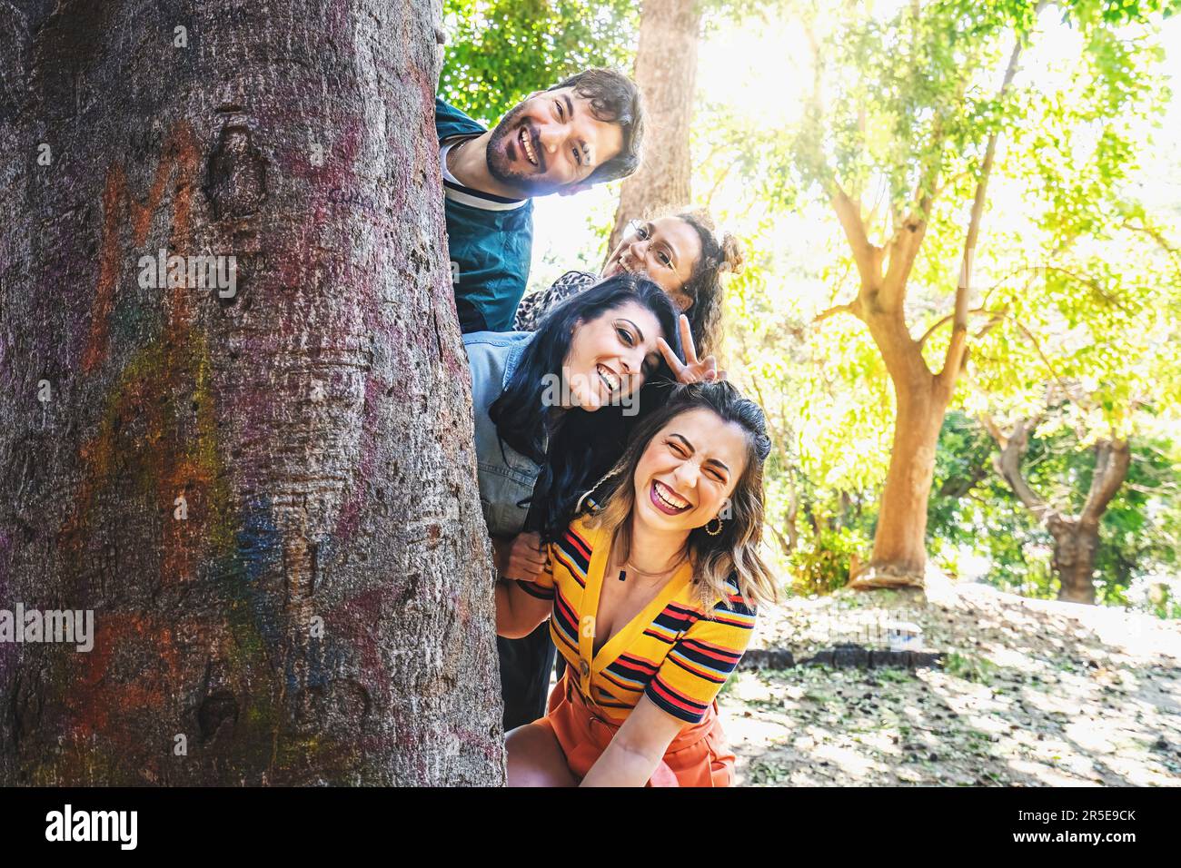 Eine Gruppe verschiedener Freunde macht ein lustiges Fotoshooting im Park. Sie verstecken ihre Köpfe spielerisch hinter einem großen Baum und fangen ehrliche Momente von laug ein Stockfoto