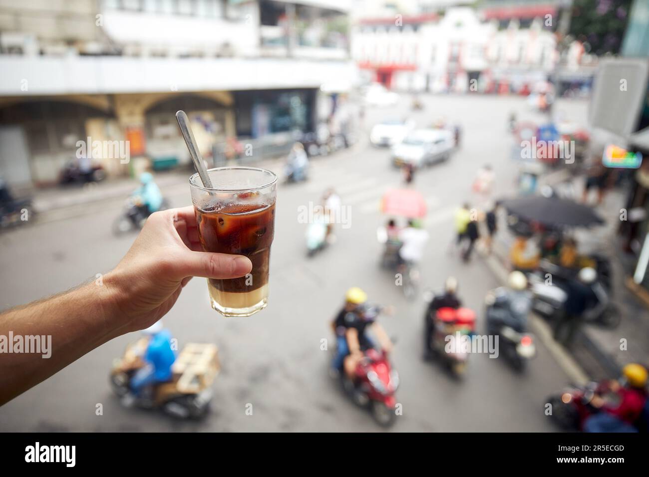Halten Sie ein Glas vietnamesischen Eiskaffee mit Kondensmilch in der Hand. Mann, der Kaffee über der belebten Straße der Altstadt in Hanoi in Vietnam trinkt. Stockfoto