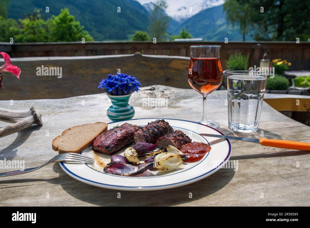Fertiggerichte - gegrilltes Chamoisfleisch mit roten und weißen Zwiebeln von einem Holzkohlegrill im Freien und ein Glas Wein und Wasser auf dem Balkon bei Sonnenschein Stockfoto