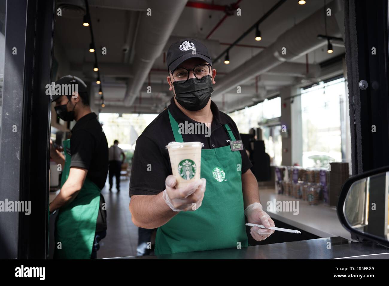 Starbucks Mitarbeiter geben Befehle mit fröhlichem Gesichtsausdruck und tragen Gesichtsmasken am Drive-in. Eisgekühlter weißer Mokka-Kaffee. Stockfoto