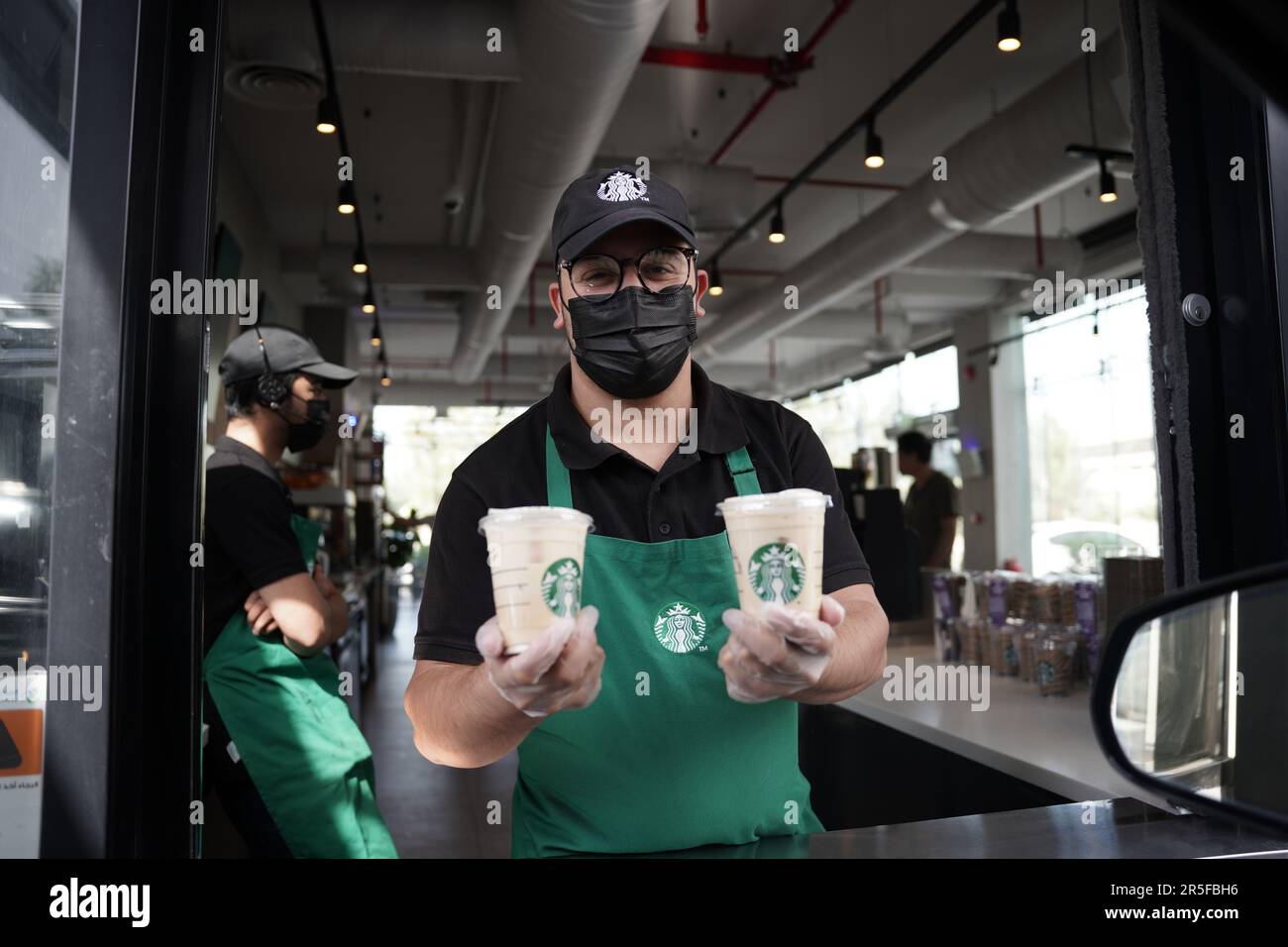 Starbucks Mitarbeiter geben Befehle mit fröhlichem Gesichtsausdruck und tragen Gesichtsmasken am Drive-in. Eisgekühlter weißer Mokka-Kaffee. Stockfoto