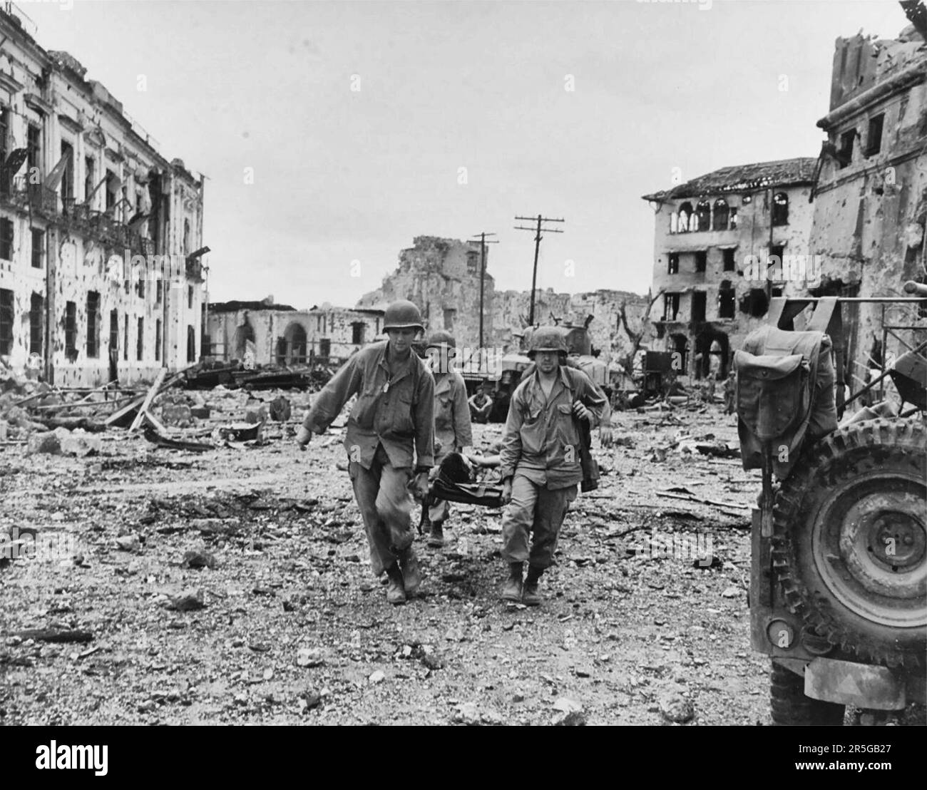 Die Trage-Party bringt einen verwundeten US-Soldaten nach einem Angriff von US-Truppen zur Befreiung philippinischer Gefangener in der ummauerten Stadt am 23. Februar 1945 heraus. Beachten Sie zerstörte Gebäude. Stockfoto