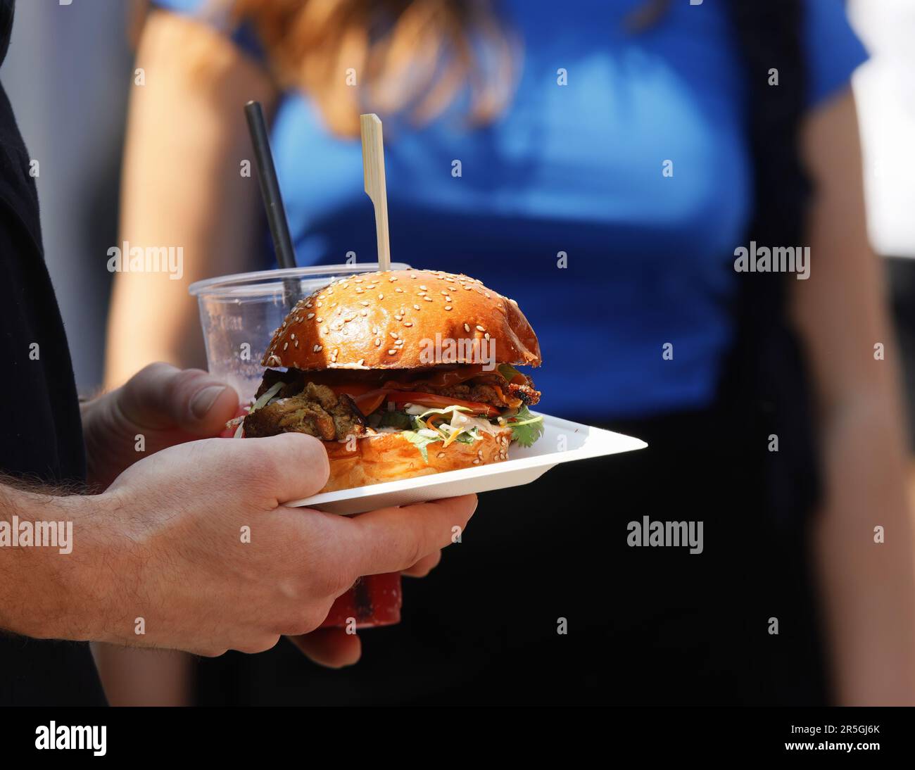 Ein Besucher des Bauernmarktes hält einen frischen Hamburger und einen Plastikbecher mit einem Getränk. Stockfoto