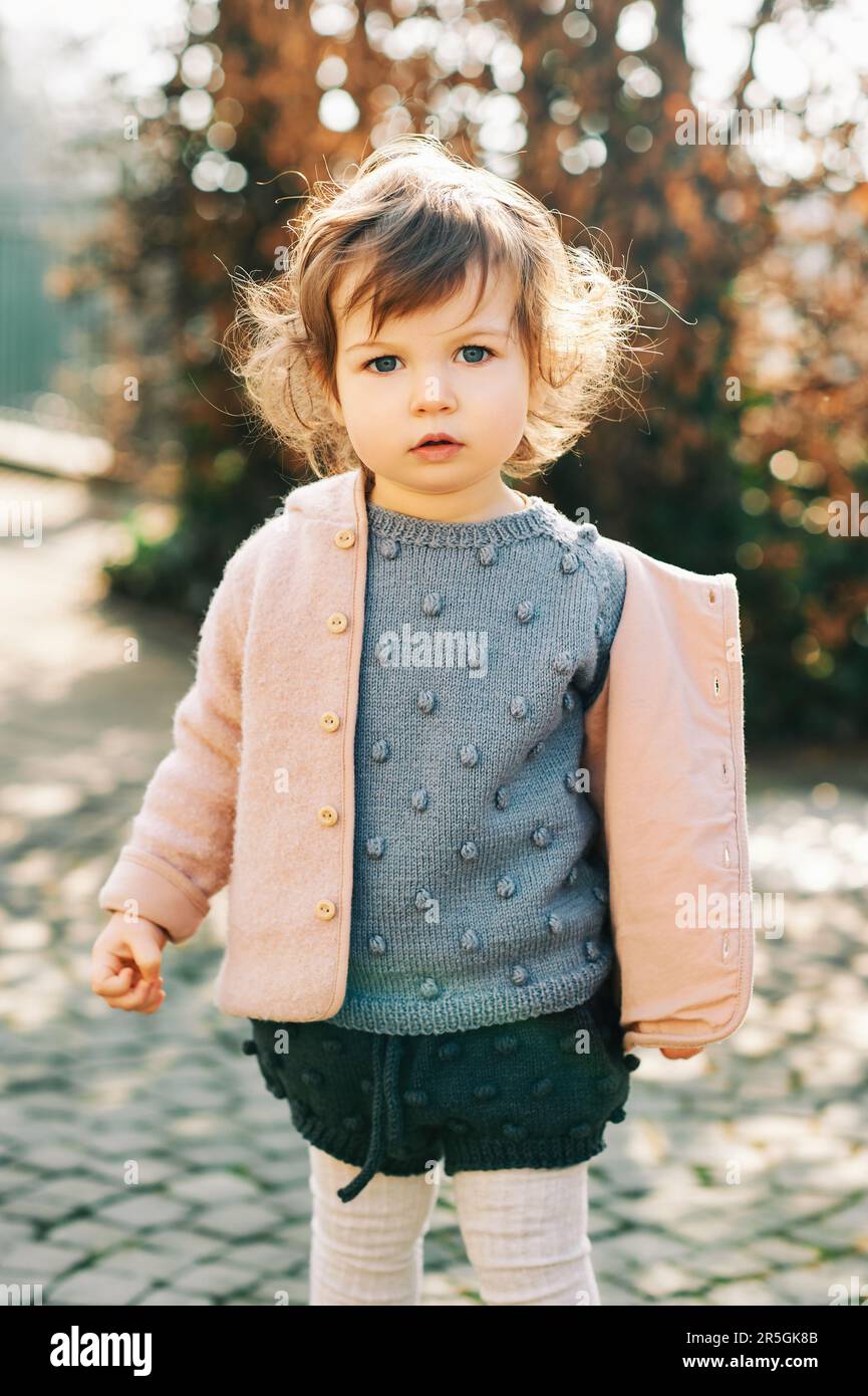 Außenporträt eines niedlichen Kleinkindes (1 -1,5 Jahre), das bei kaltem Wetter einen blauen Pullover und eine rosa Jacke trägt Stockfoto