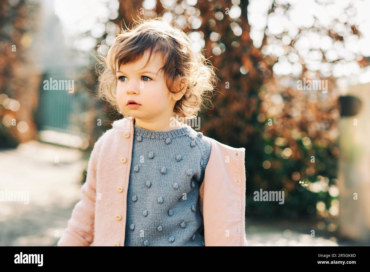 Außenporträt eines niedlichen Kleinkindes (1 -1,5 Jahre), das bei kaltem Wetter einen blauen Pullover und eine rosa Jacke trägt Stockfoto