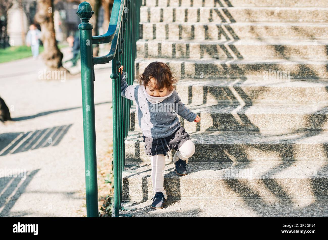 Außenporträt eines niedlichen Kleinkindes, das die Treppe hinaufgeht, ein Kind von 1 bis 2 Jahren, das Treppen auf der Straße der Stadt hochklettert und einen gestrickten Pullov trägt Stockfoto