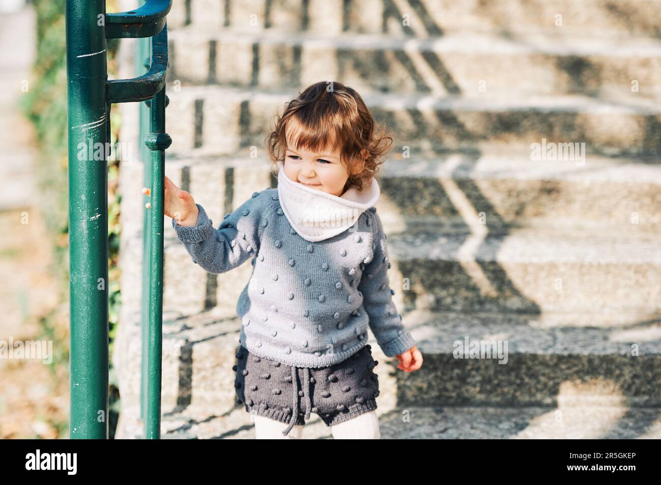 Außenporträt eines niedlichen Kleinkindes, das die Treppe hinaufgeht, ein Kind von 1 bis 2 Jahren, das Treppen auf der Straße der Stadt hochklettert und einen gestrickten Pullov trägt Stockfoto