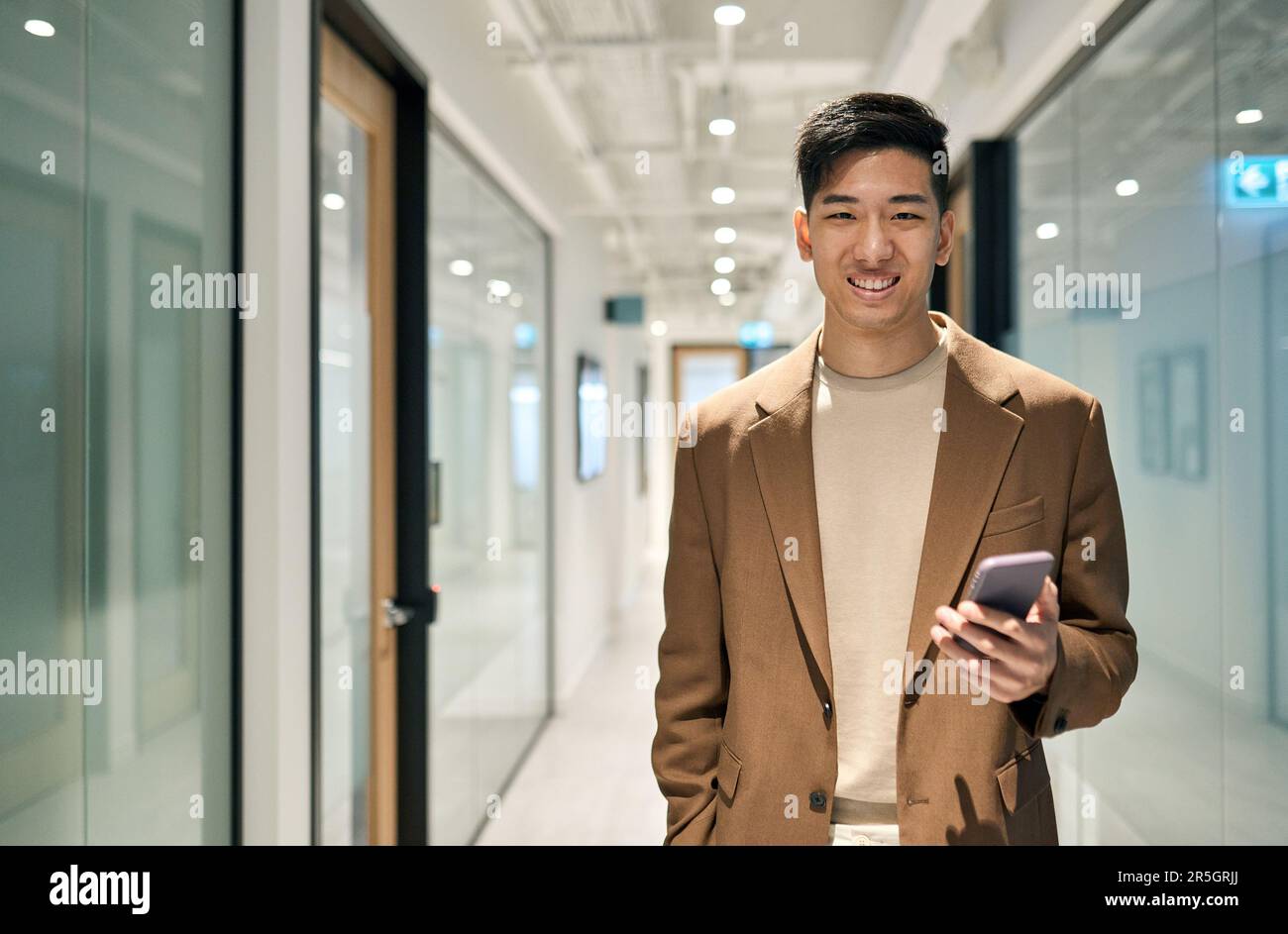 Junger, glücklicher asiatischer Geschäftsmann, der im Flur steht und ein Telefon hält. Stockfoto