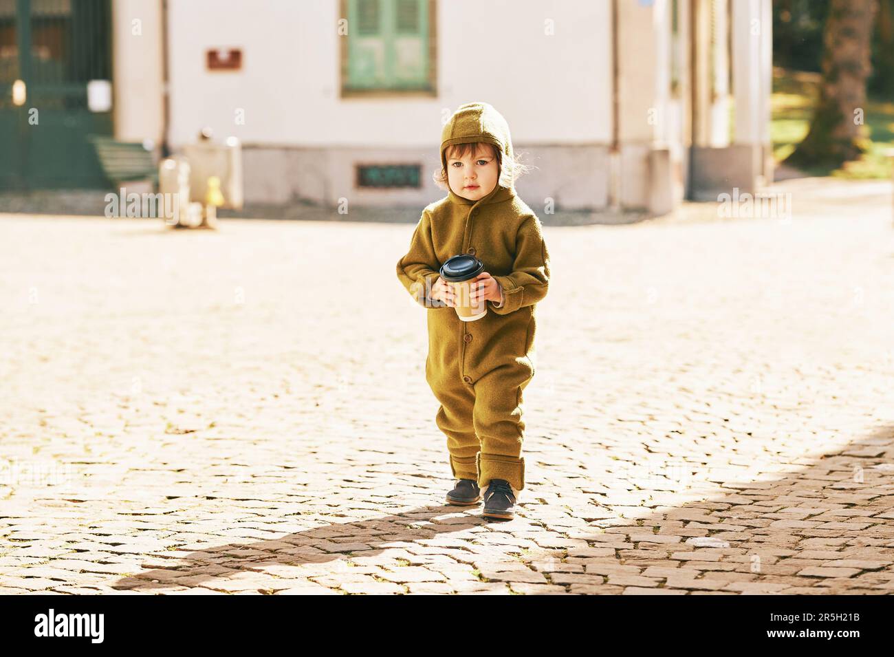 Außenporträt eines niedlichen Kleinkindes, das die Straße entlang läuft, einen olivfarbenen, warmen Overall trägt und eine Tasse Kaffee zum Mitnehmen hält Stockfoto