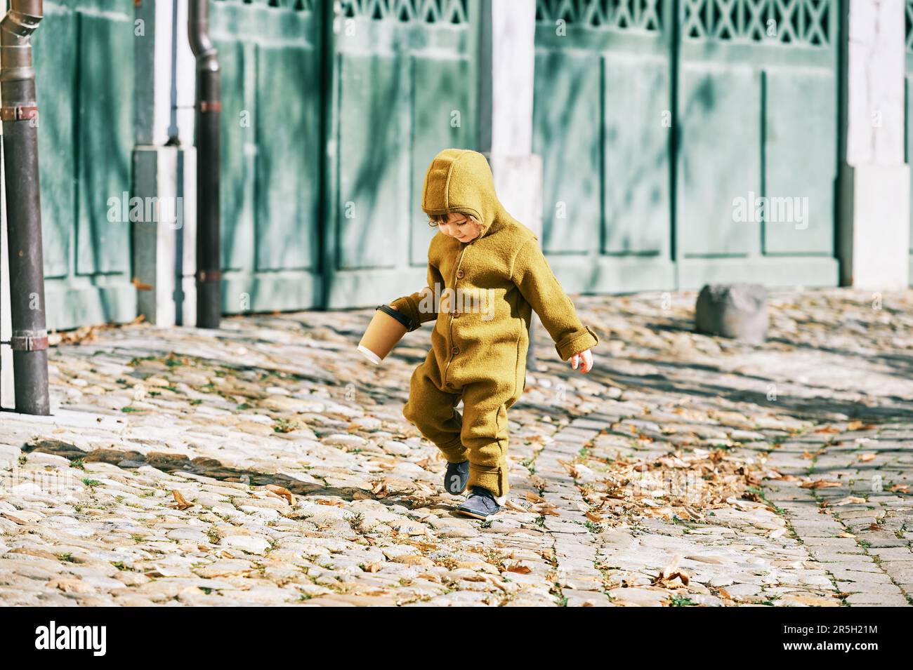 Außenporträt eines niedlichen Kleinkindes, das die Straße entlang läuft, einen olivfarbenen, warmen Overall trägt und eine Tasse Kaffee zum Mitnehmen hält Stockfoto