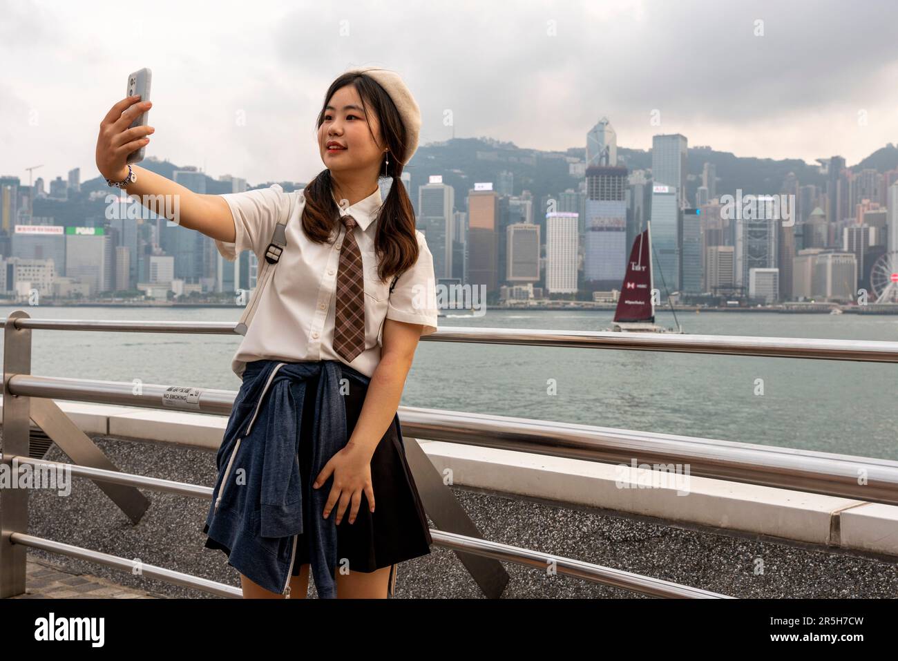 Eine junge Chinesin nimmt Ein Selfie auf der Avenue of Stars mit Hong Kong Island als Kulisse, Kowloon, Hongkong, China. Stockfoto