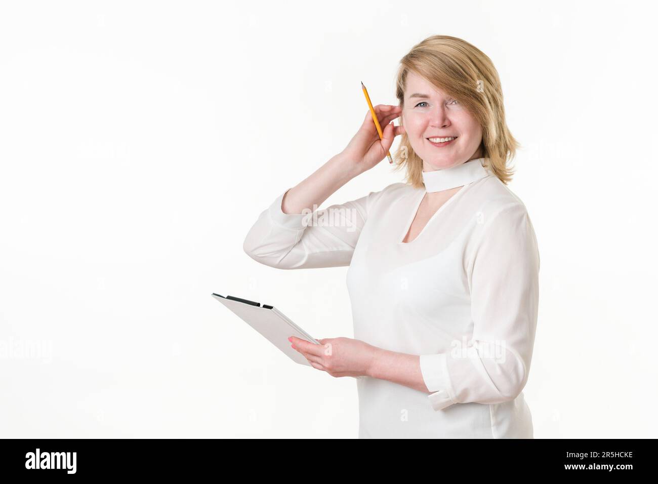 Eine lächelnde Lehrerin glättet blondes Haar mit der Hand mit einem Bleistift. Glückliche Frau, die das Klemmbrett hält und in die Kamera schaut. Blonde Frau Stockfoto