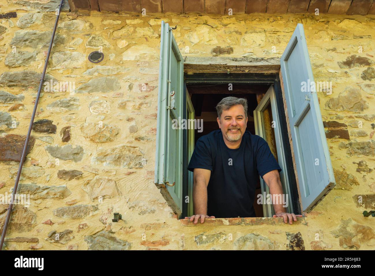 Blick von außen auf den Mann, der aus dem Fenster aus dem zweiten Stock in einem alten Steinhaus mit Fensterläden geneigt war Stockfoto