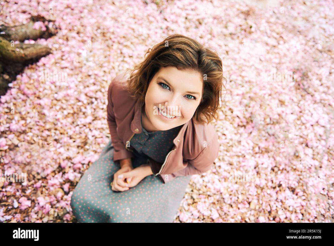 Frühlingsporträt im Freien mit einer lächelnden glücklichen Frau, die in die Kamera schaut, rosa Blütenblätter auf dem Hintergrund blüht und eine braune Lederjacke trägt Stockfoto