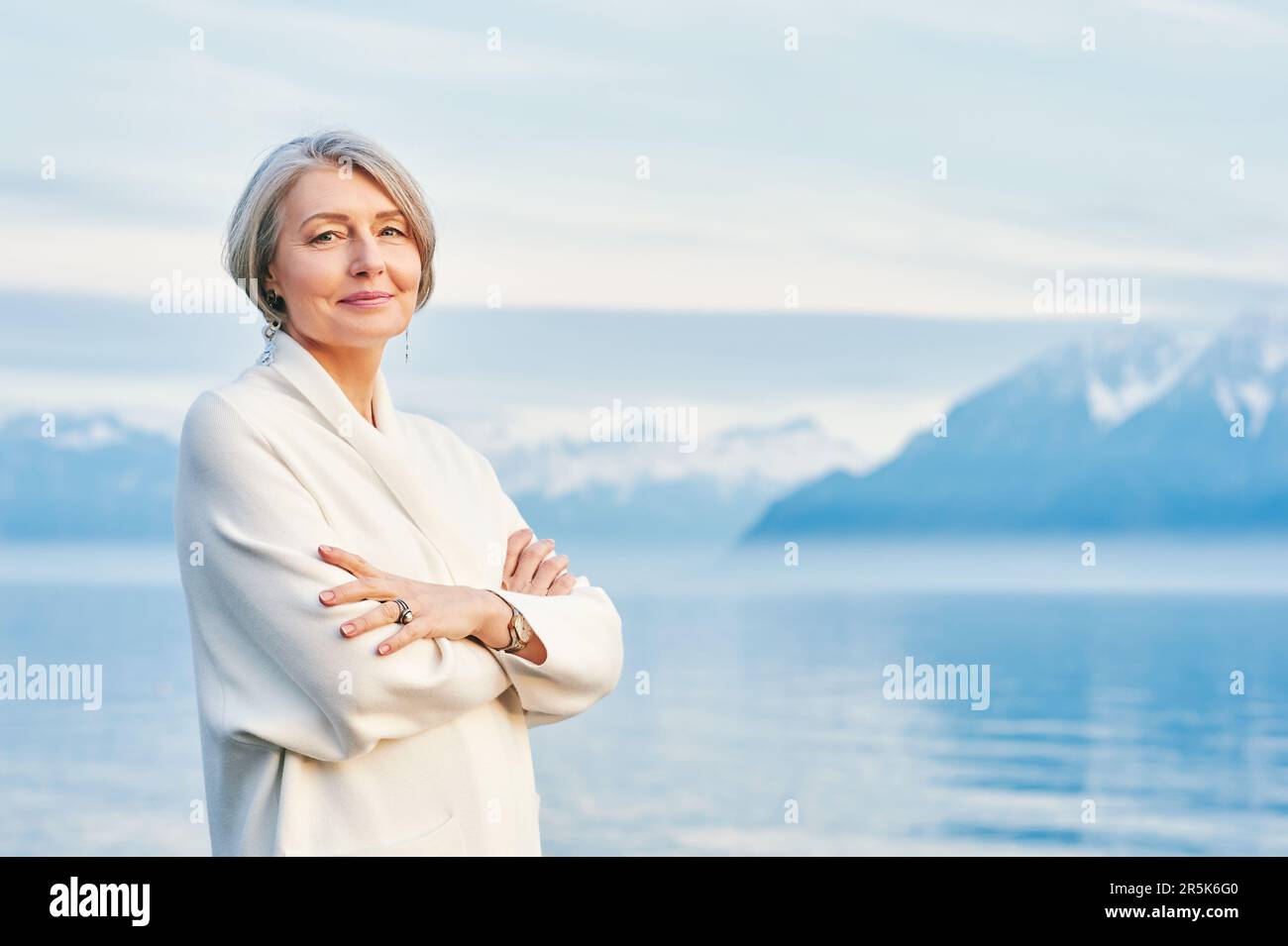 Außenporträt einer wunderschönen Frau mittleren Alters, die sich am Wintersee posiert und einen weißen Mantel trägt Stockfoto