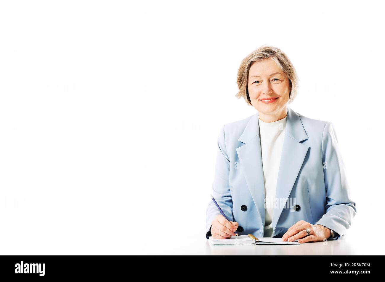Studioporträt einer Frau mittleren Alters, die auf weißem Hintergrund posiert, am Schreibtisch sitzt und sich Notizen macht Stockfoto