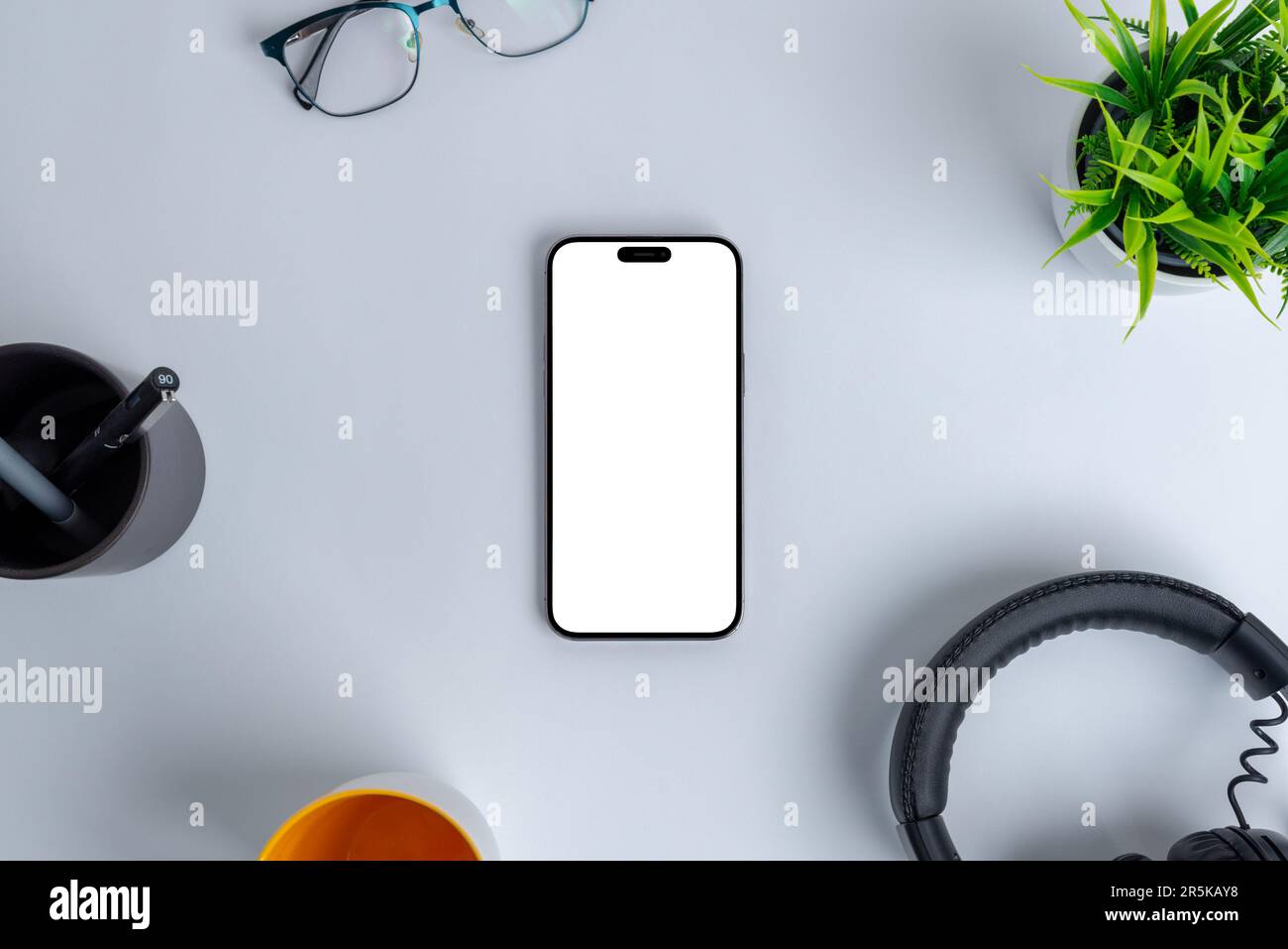 Moderne Arbeitsumgebung mit einem leeren Telefonbildschirm, der zur Nachahmung ausgelegt ist und sich zwischen Büroutensilien wie Headset, Pflanze, Brille, Kugelschreiber und Kaffee befindet Stockfoto