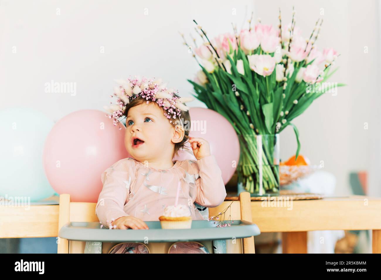 Ein bezauberndes Mädchen, das auf einem Stuhl sitzt, Cupcake mit einer Kerze ansieht, erste Geburtstagsparty Stockfoto