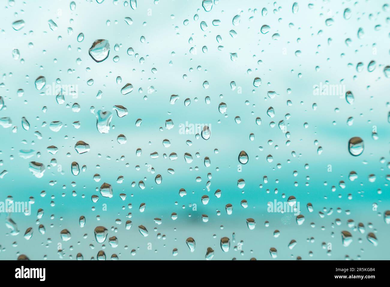 Tropfen auf ein Fensterglas mit verschwommenem blauem Hintergrund Nahaufnahme, Regenmakro Stockfoto