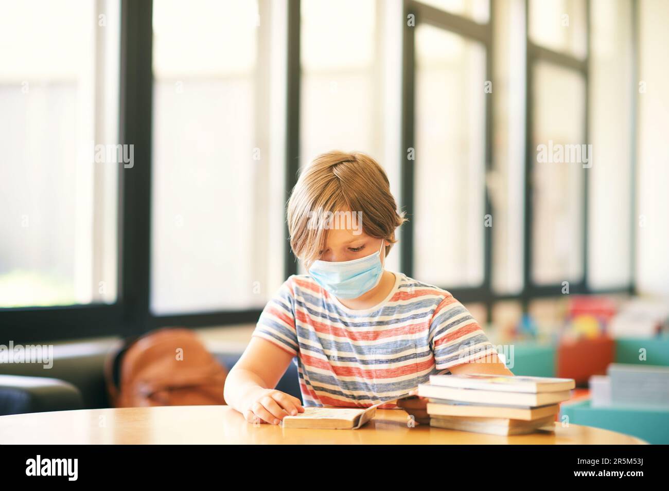 Ein kleiner Junge sitzt in einem Klassenzimmer oder einer Bibliothek, trägt eine Gesichtsmaske, hält ein Buch, zurück zur Schule Konzept Stockfoto