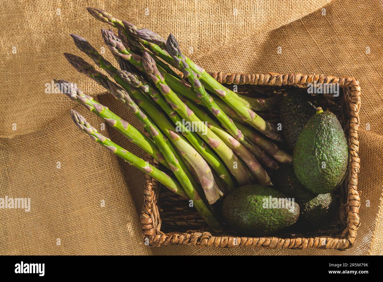 Frischer Bio-Spargel und Avocados in einem Wicker-Korb aus der Nähe auf rustikalem Hintergrund, Blick von oben Stockfoto