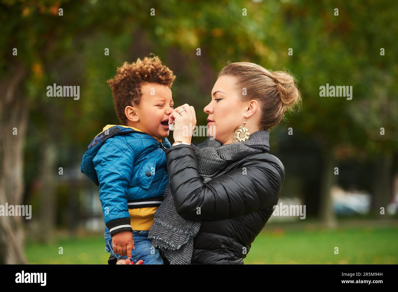 Junge Mutter hält weinendes Kleinkind in der Hand, Elternteil im Park mit traurigem Kind, gemischte Rennfamilie Stockfoto