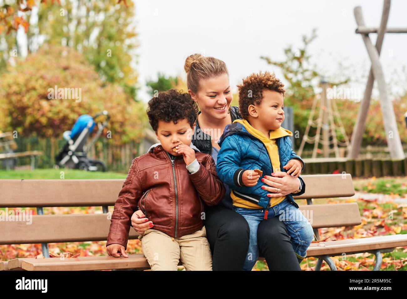 Außenporträt einer glücklichen jungen Mutter mit zwei reizenden Söhnen, einer Familie, die einen schönen Herbsttag im öffentlichen Park genießt, einer Familie mit gemischten Rassen. Kinder essen Snacks, Wiedereinstieg Stockfoto