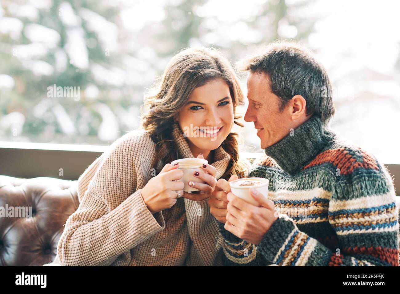 Ein Paar mittleren Alters, das sich auf der Ledercouch ausruht, Kaffee trinkt, warme, gemütliche Pullover trägt Stockfoto