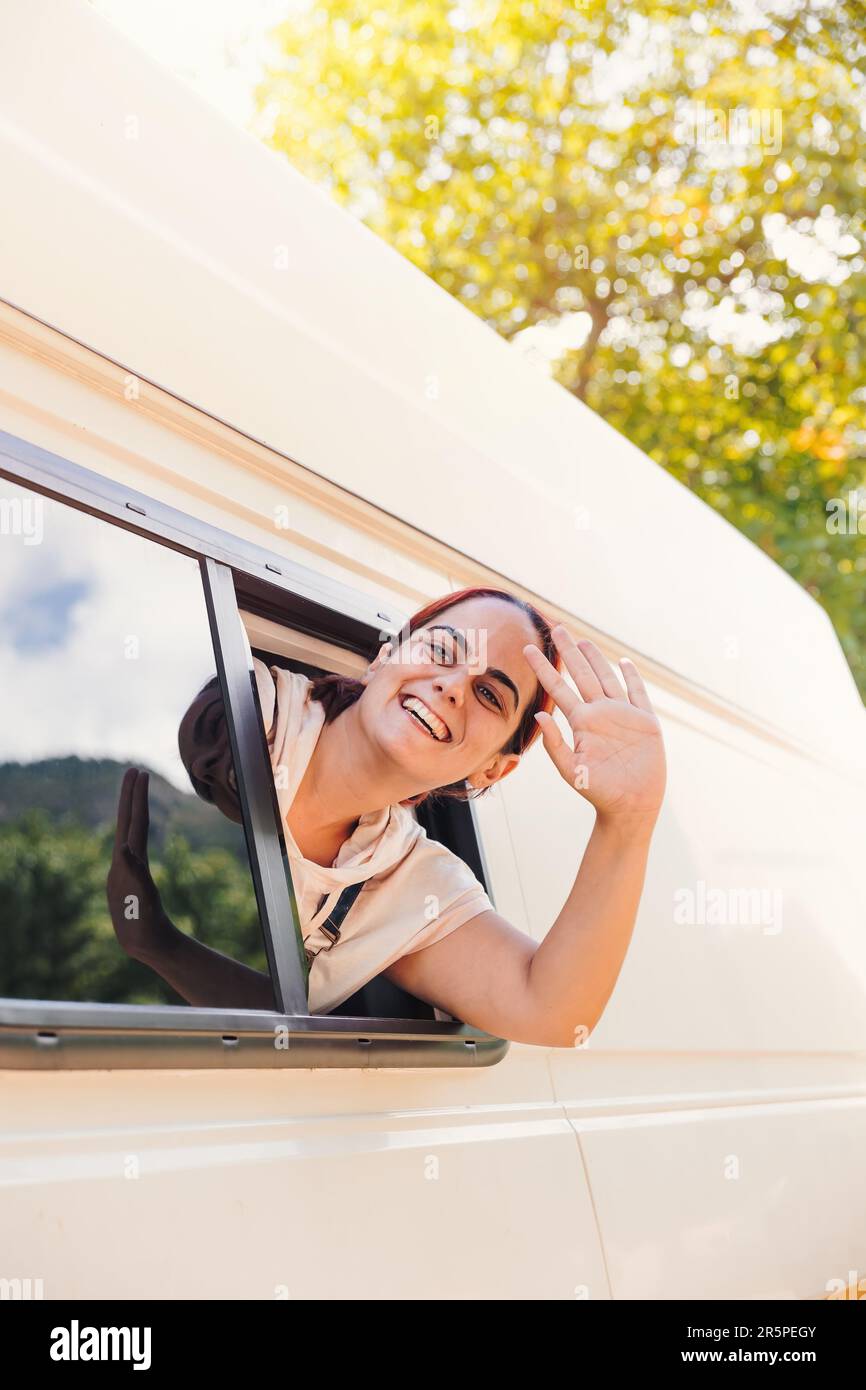 Junge Frau winkt durch das Fenster ihres Wohnwagens. Roadtrip im Minibus und Sommerabenteuer im Freien. Nomad Lifestyle-Konzept Stockfoto