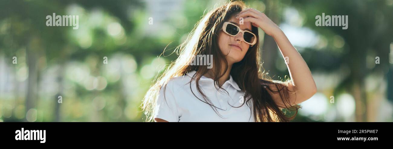 Sommeratmosphäre, hübsche Frau, die lange braune Haare anpasst und in weißem Polo-Shirt und Sonnenbrille auf der Straße in Miami steht, verschwommener Hintergrund, Stockfoto