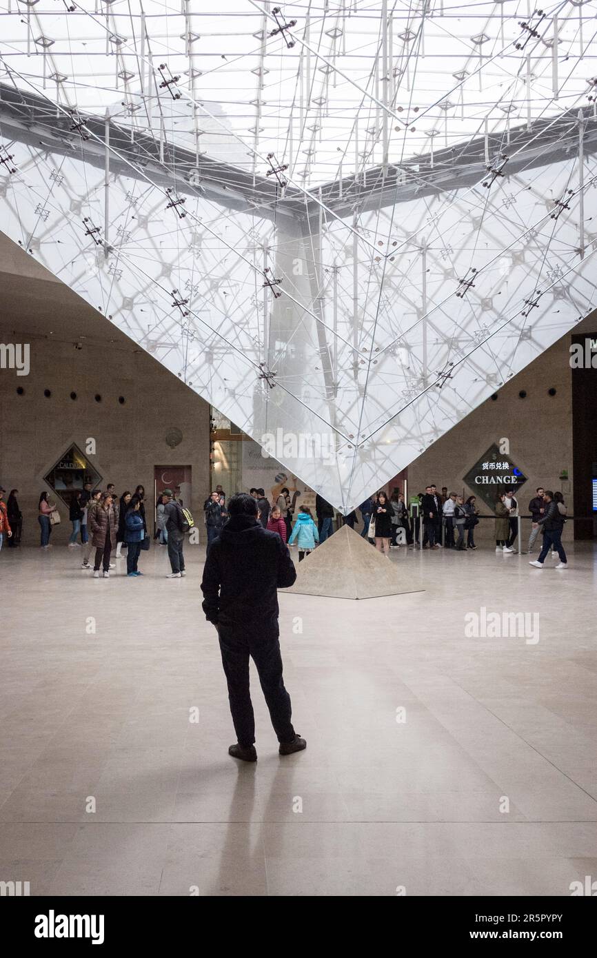 Die Menschen in Paris, Frankreich, versammeln sich um die fesselnde umgekehrte Pyramide, die sich im bekannten Einkaufszentrum „Carrousel du Louvre“ befindet, während sich die Menschen anstellen, um das Museum im Hintergrund zu betreten. Stockfoto